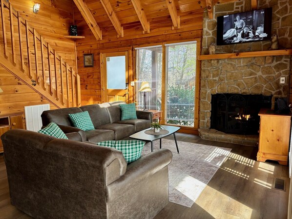 A wooden interior with a fireplace, a television, and a sofa with checkered cushions. - Pet friendly, hot tub, cozy log cabin sleeps 7! (Sevierville)