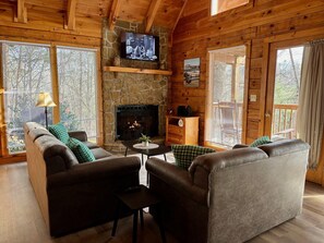 A cozy cabin interior with a stone fireplace, wooden walls, a television mounted above it, and a comfortable seating area with sofas and a coffee table.