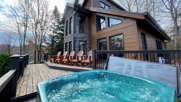 A modern two-story house with a wooden exterior, a deck with lounge chairs, and a hot tub.