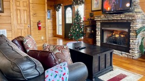A cozy log cabin interior with a fireplace, a television, a Christmas tree, and a leather sofa.