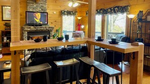 A wooden cabin interior with a stone fireplace, a television, and a bar area with stools.