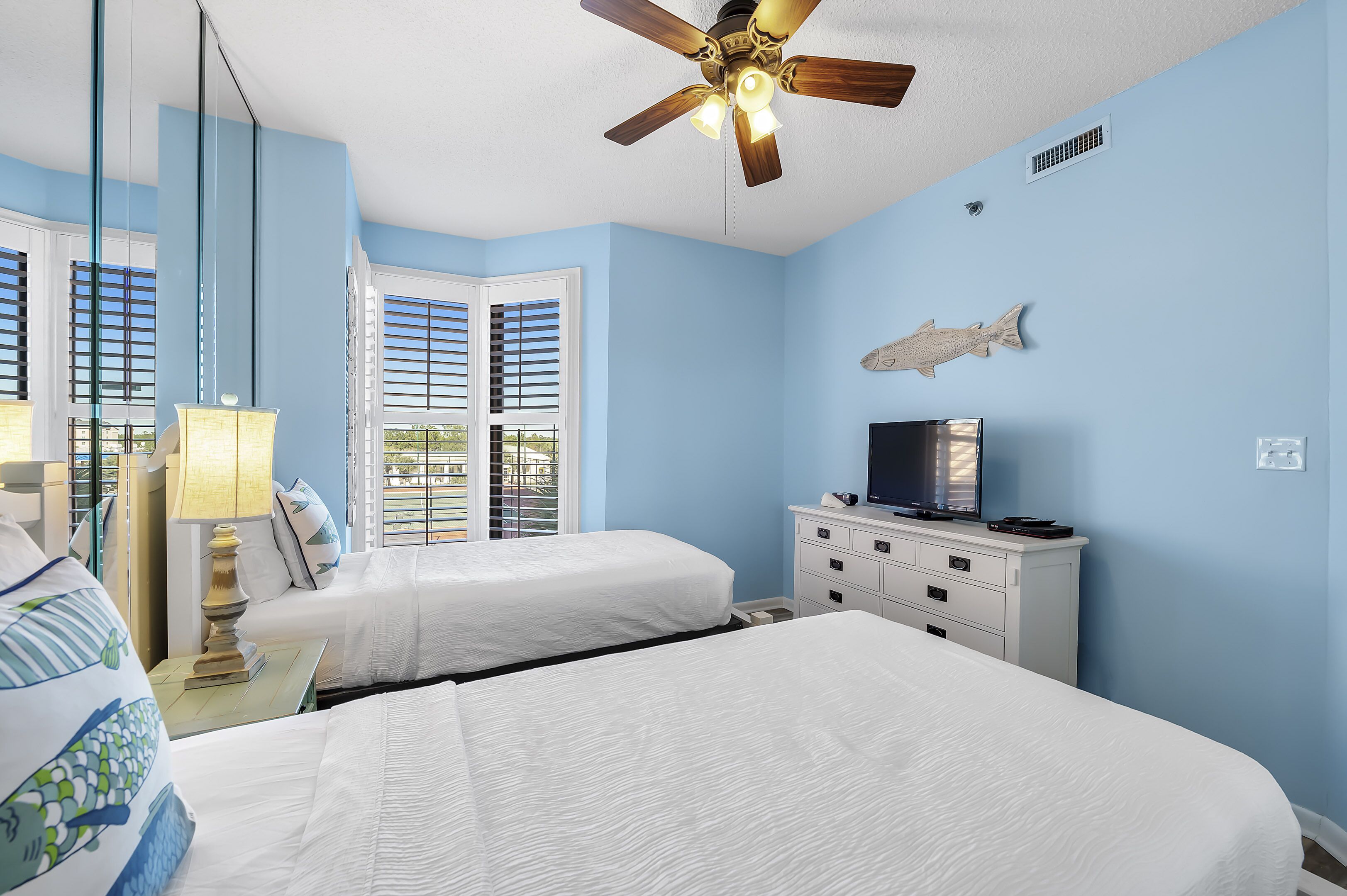 A hotel room with two beds, a ceiling fan, a TV on a dresser, and a fish decoration on the wall.