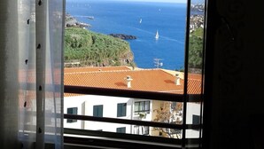 Blick aus einem Fenster mit Vorhang auf eine Küstenlandschaft mit Gebäuden und dem Meer.