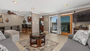 A living room with a sofa, a coffee table, a dining area with chairs, a kitchen in the background, and a large sliding glass door leading to a balcony. - Beachfront Condo with Heated Pool and Hot Tub | 311B (Oceanside)