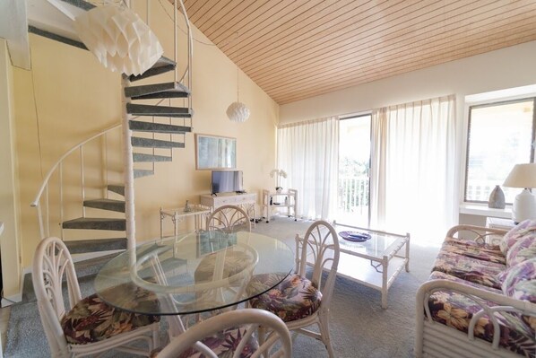A spacious living area with a glass-top dining table, white wicker furniture, a staircase, and large windows.