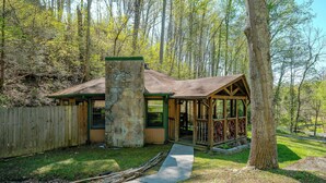 A rustic cabin with a stone chimney, wooden deck, and a view of the surrounding forest.