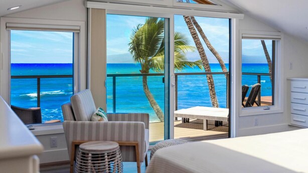 Ein Schlafzimmer mit großem Fenster und Meerblick, einem Balkon mit Tisch und Stühlen sowie Blick auf Palmen.