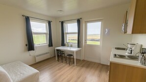 A small, bright room with a single bed, a table with two chairs, a kitchen area with a stove and sink, and large windows with curtains.