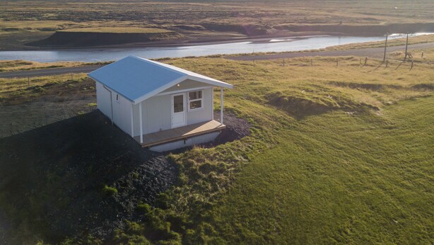 A small white cabin with a blue roof, situated on a grassy hill overlooking a body of water and a cloudy sky.