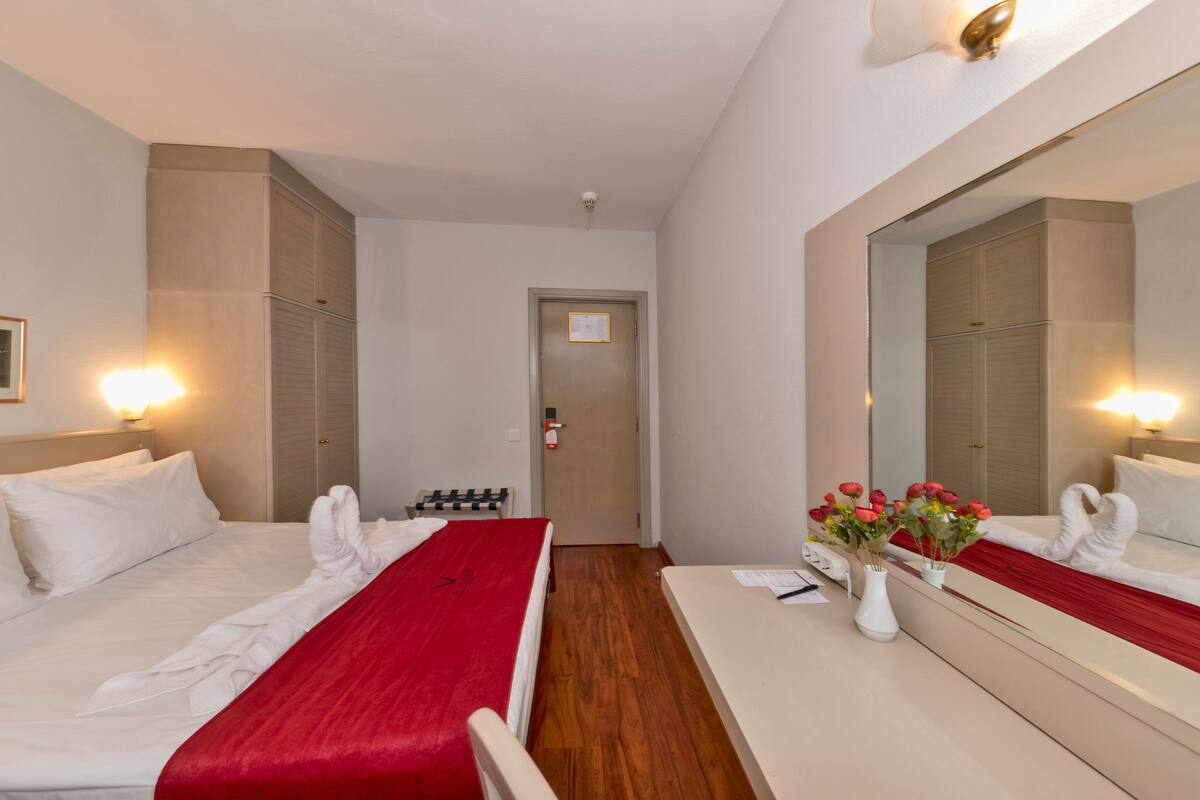 Habitación de hotel con dos camas, piso de madera, un espejo grande y un jarrón con flores.