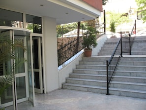 A set of stone steps leading up to a building entrance, with a metal railing on the right and a glass door on the left.
