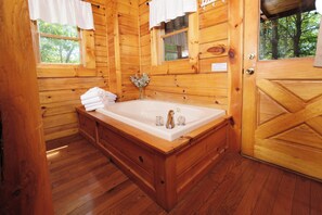 Un baño de cabaña de madera con una bañera de madera, dos ventanas con cortinas blancas y una puerta.