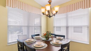 A dining room with a wooden table set for six, surrounded by chairs, with a chandelier above and striped window valances. - Modern Villa w/ South-Facing Pool, Spa & Game Room Near Disney (Kissimmee)