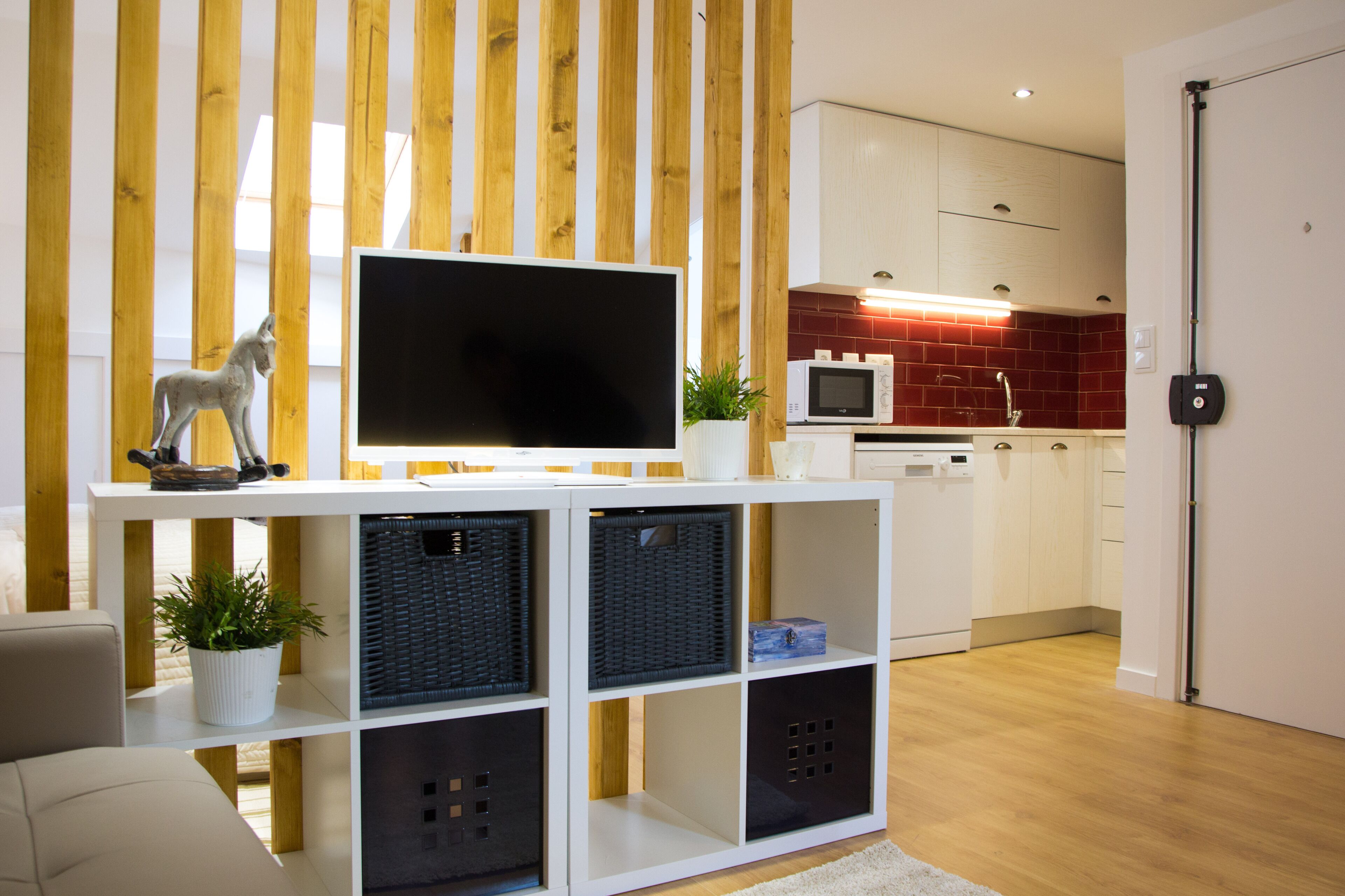 A modern living room with a flat-screen TV on a white console, black baskets, and a potted plant. A wooden slat wall and a kitchen with red tiles are visible in the background.