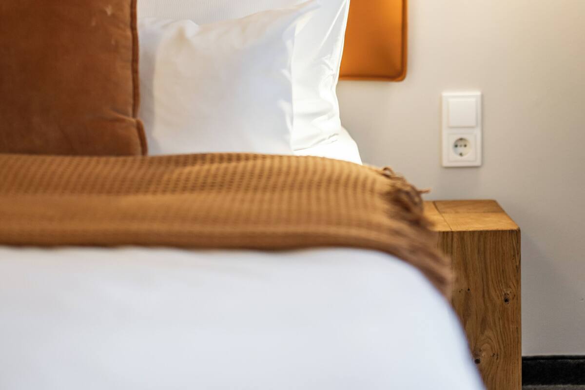 A neatly made bed with white and brown bedding, a wooden nightstand, and a wall-mounted light fixture.