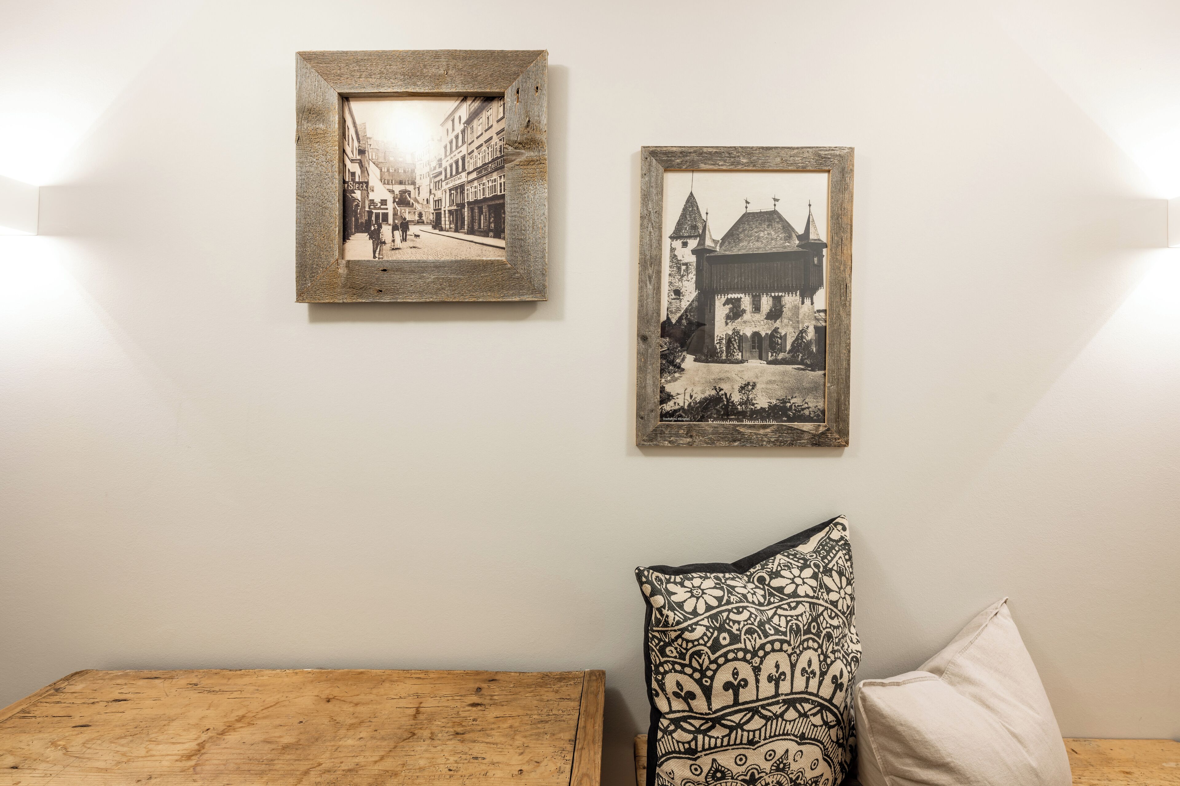 Deux photographies en noir et blanc encadrées, accrochées au mur, un coussin à motifs et un banc en bois.