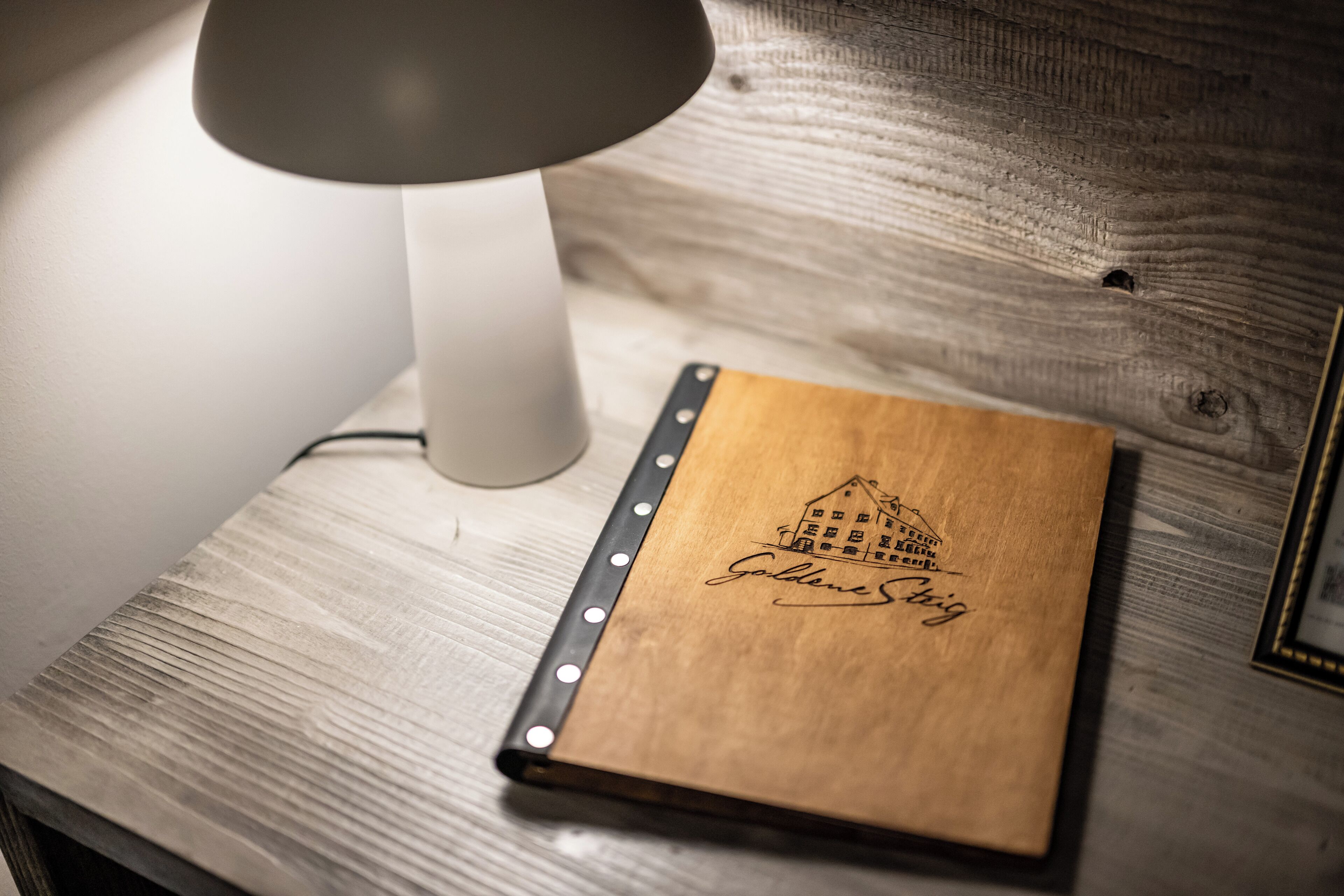 Un cuaderno de madera con un grabado de una casa y el nombre "GoldenSieg" sobre una superficie de madera, junto a una lámpara blanca.