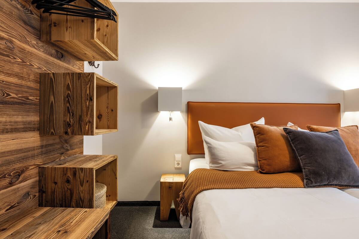 A hotel room with a wooden wall unit, a bed with multiple pillows, a nightstand, and a wall-mounted lamp.
