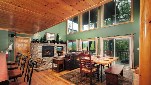 Un salon chaleureux avec une cheminée, un plancher en bois, un coin repas et de grandes fenêtres donnant sur l’extérieur.