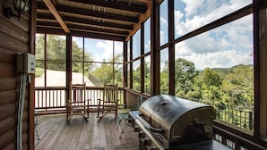 A wooden deck with a grill, chairs, and a view of a forested area.