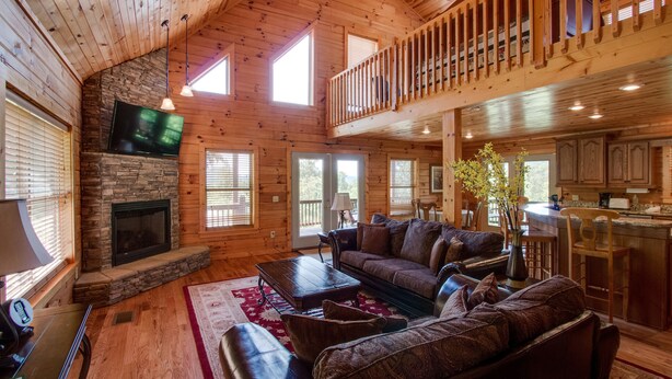 A spacious living room with a stone fireplace, a large flat-screen TV, a wooden ceiling, and a balcony with a railing.