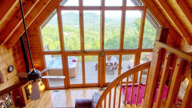 Una cabaña de madera con un gran ventanal que da a una terraza. En su interior, hay un ventilador de techo, una mesita y una silla.