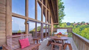 Una terraza de madera con banco y mesa de picnic, ventanas amplias y vistas al verdor.