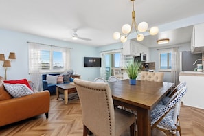 Un salon moderne avec une table à manger en bois, des chaises recouvertes d’un tissu beige, un canapé bleu et un ventilateur de plafond.