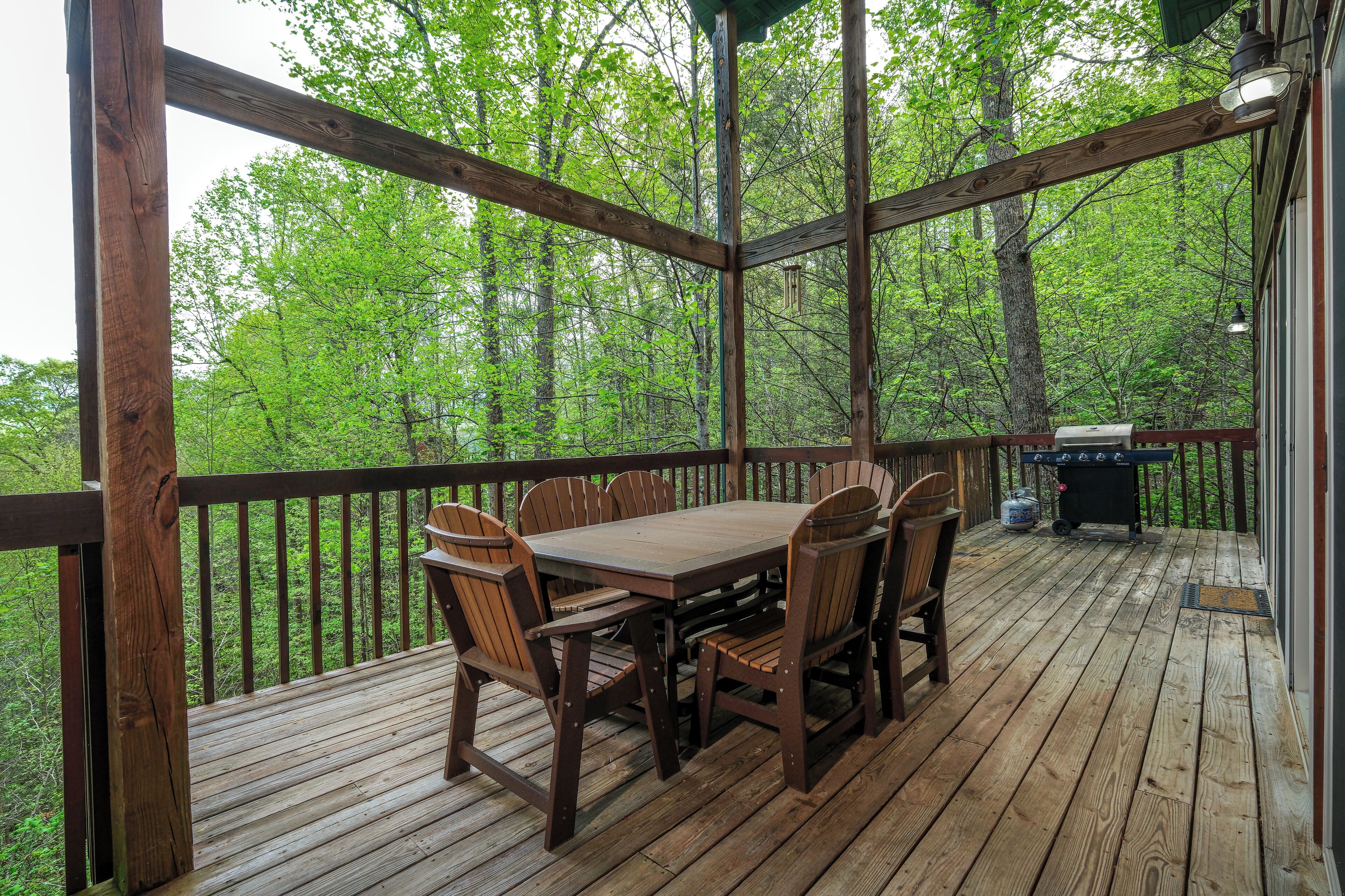 A wooden deck with a table and chairs, surrounded by a railing, overlooking a forested area.