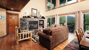 A cozy living room with a stone fireplace, wooden furniture, and large windows overlooking a deck. - Mountain views, pet friendly, game rooms, 3 king bedrooms, and 2 game rooms! (Sevierville)
