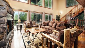 A spacious living room with a stone fireplace, large windows, and a wooden staircase. - Mountain views, pet friendly, game rooms, 3 king bedrooms, and 2 game rooms! (Sevierville)
