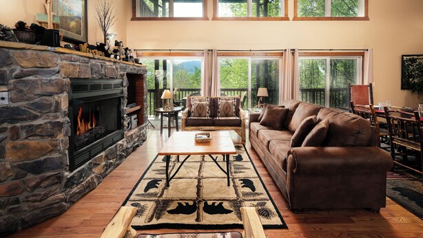 A cozy living room with a stone fireplace, a brown leather sofa, and a large window overlooking a forest. - Mountain views, pet friendly, game rooms, 3 king bedrooms, and 2 game rooms! (Sevierville)