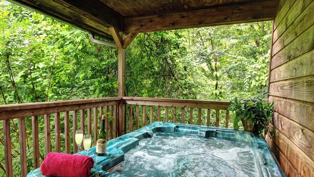 Un jacuzzi en una terraza de madera con techo de madera, rodeado de abundante vegetación.