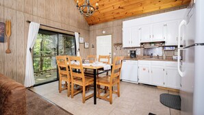 Una habitación con paneles de madera, una mesa de comedor para cuatro personas, una cocina con armarios blancos y un ventanal con vistas a un bosque.
