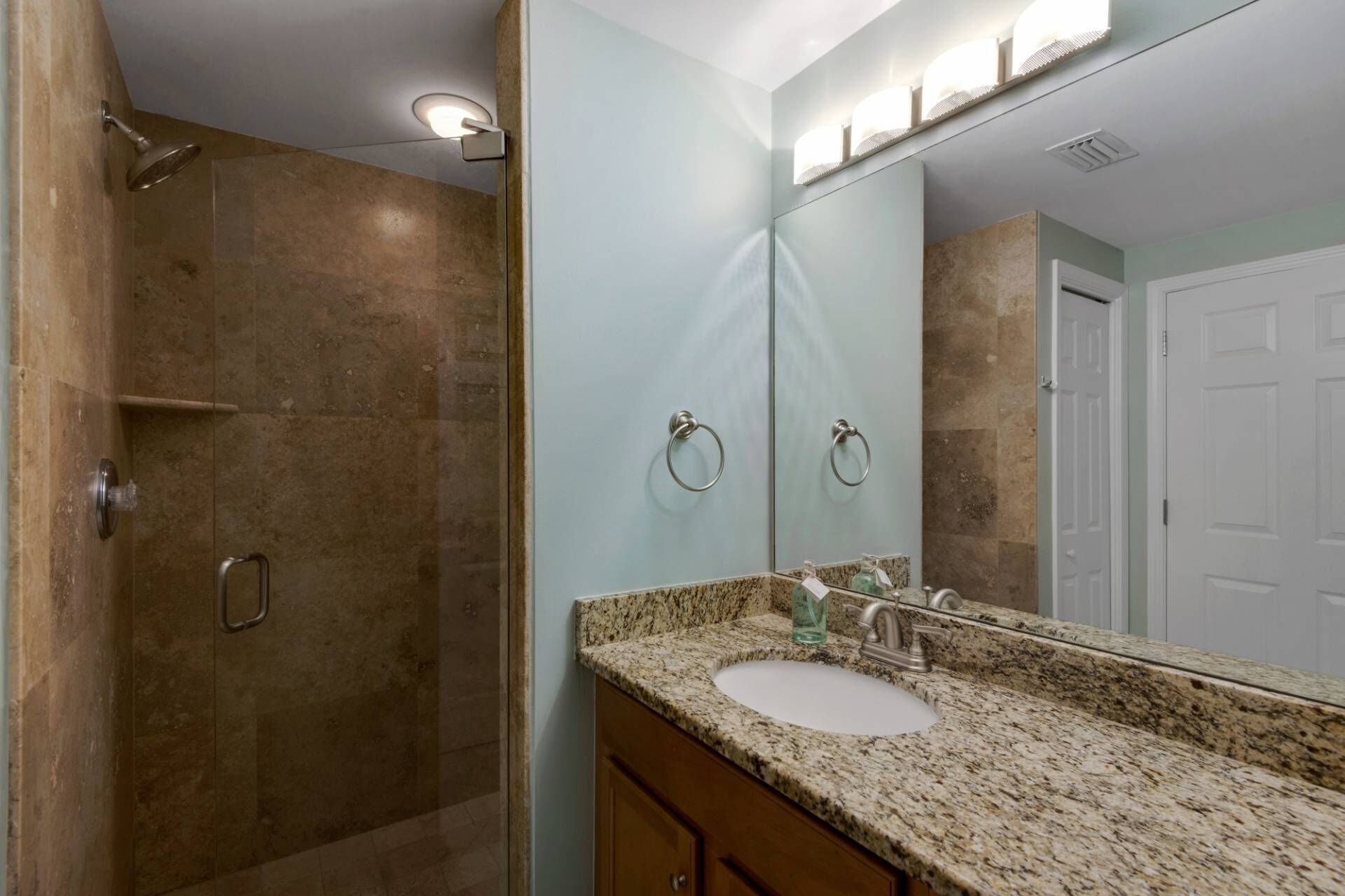 A bathroom with a granite countertop, a sink, a mirror, a shower, and a door.