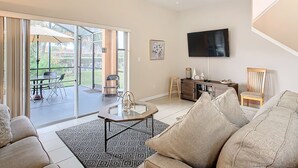 Una sala de estar con un sofá, una mesa de centro, un televisor y un ventilador de techo. Hay una puerta corrediza de vidrio que da a un patio exterior con una mesa y sillas.