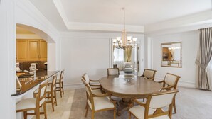 Uma sala de jantar com uma mesa redonda, seis cadeiras, um lustre e uma cozinha visível através de um arco.