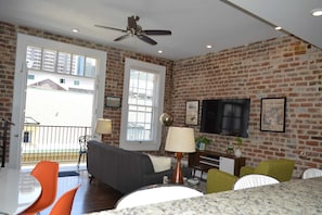 A living room with a brick wall, a ceiling fan, a flat-screen TV, and a dining area with orange chairs.
