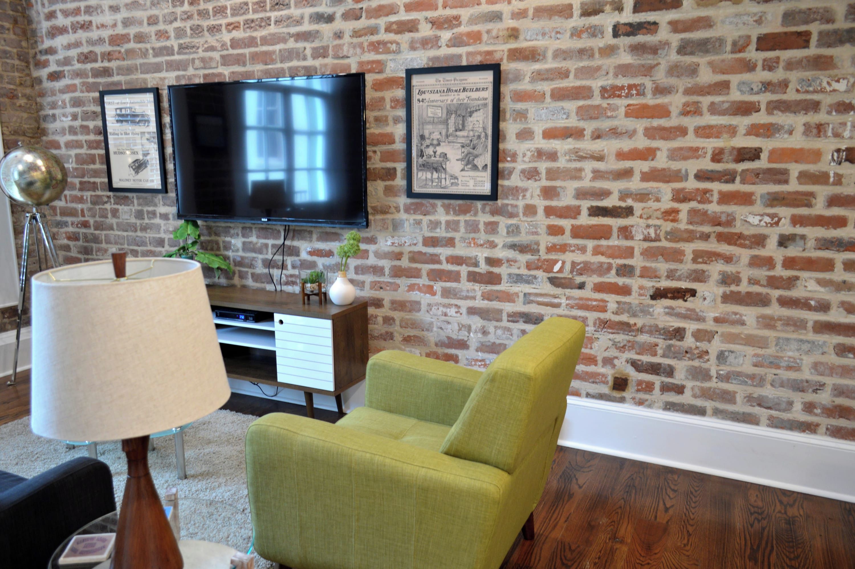 A living room with a brick wall, a flat-screen TV mounted above a wooden entertainment center, a green armchair, a floor lamp, and a small table with a vase.