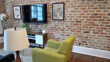 A living room with a brick wall, a flat-screen TV mounted above a wooden entertainment center, a green armchair, a floor lamp, and a small table with a vase.