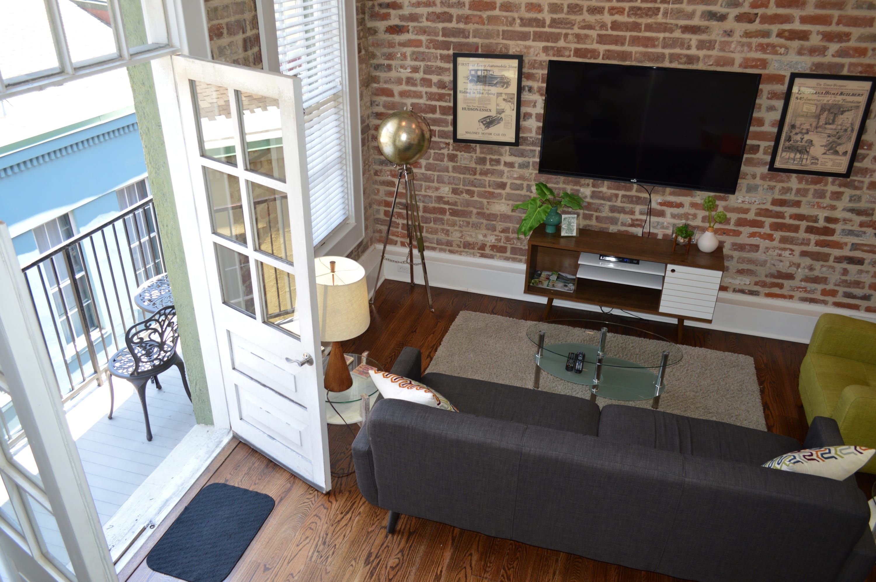 Een woonkamer met een bakstenen muur, een flatscreen televisie, een bank, een salontafel en een balkon met tuinmeubilair.