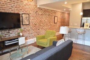 A living room with a brick wall, a flat-screen TV, a sofa, a green armchair, and a kitchen area with a refrigerator and bar stools.