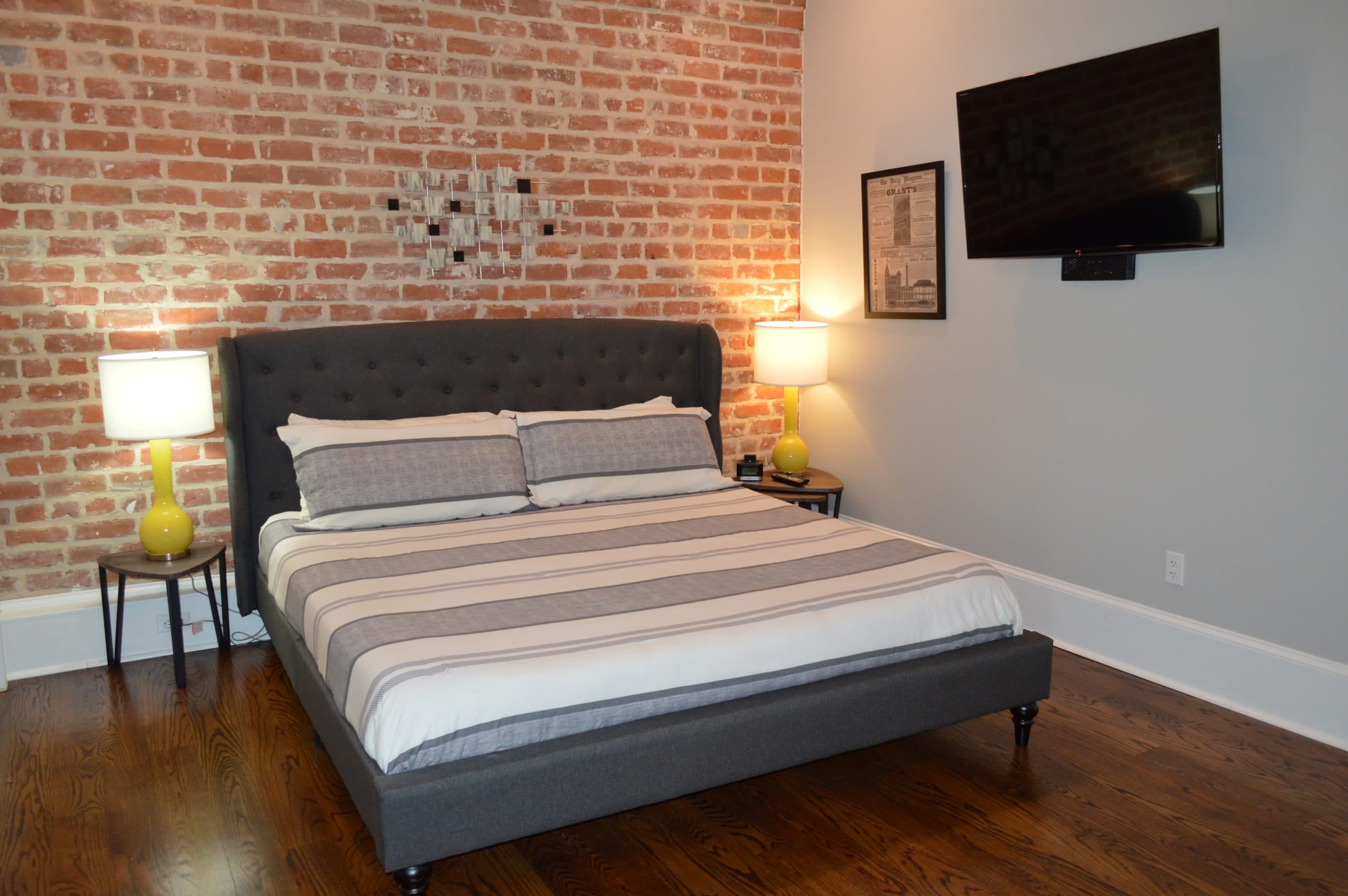 A bedroom with a brick wall, a bed with striped bedding, two bedside lamps, and a flat-screen TV mounted on the wall.