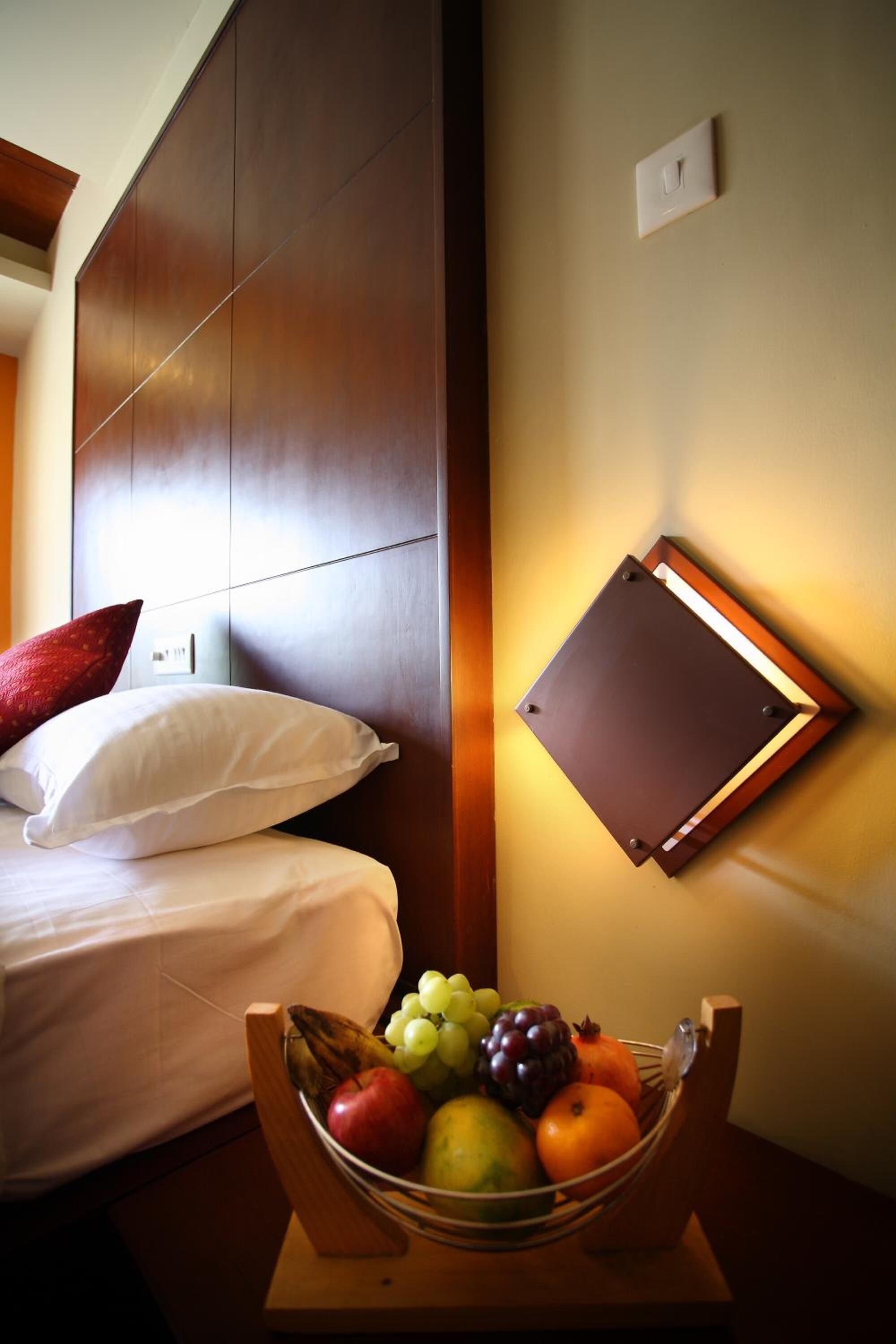 Una camera d'albergo con un letto, un comodino con una ciotola di frutta, una lampada a parete e una testiera in legno.