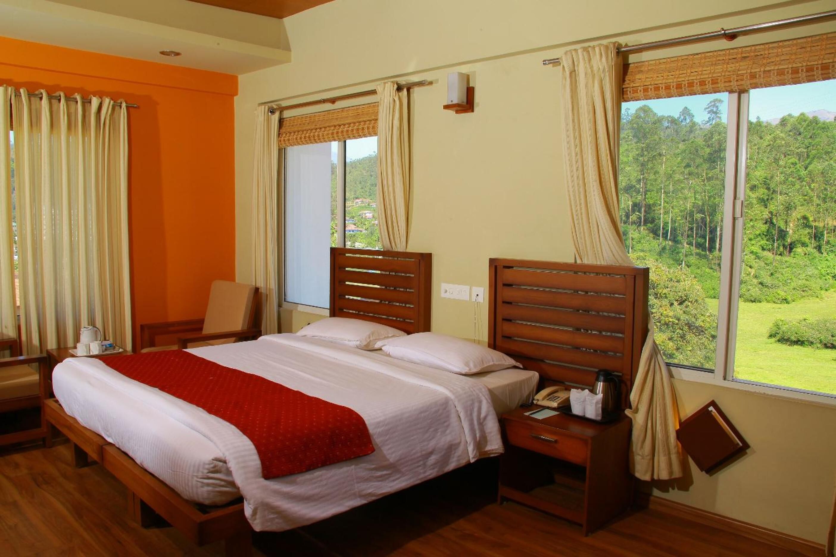 A hotel room with a large bed, wooden headboard, a bedside table with a lamp, and a view of a forest through the window.