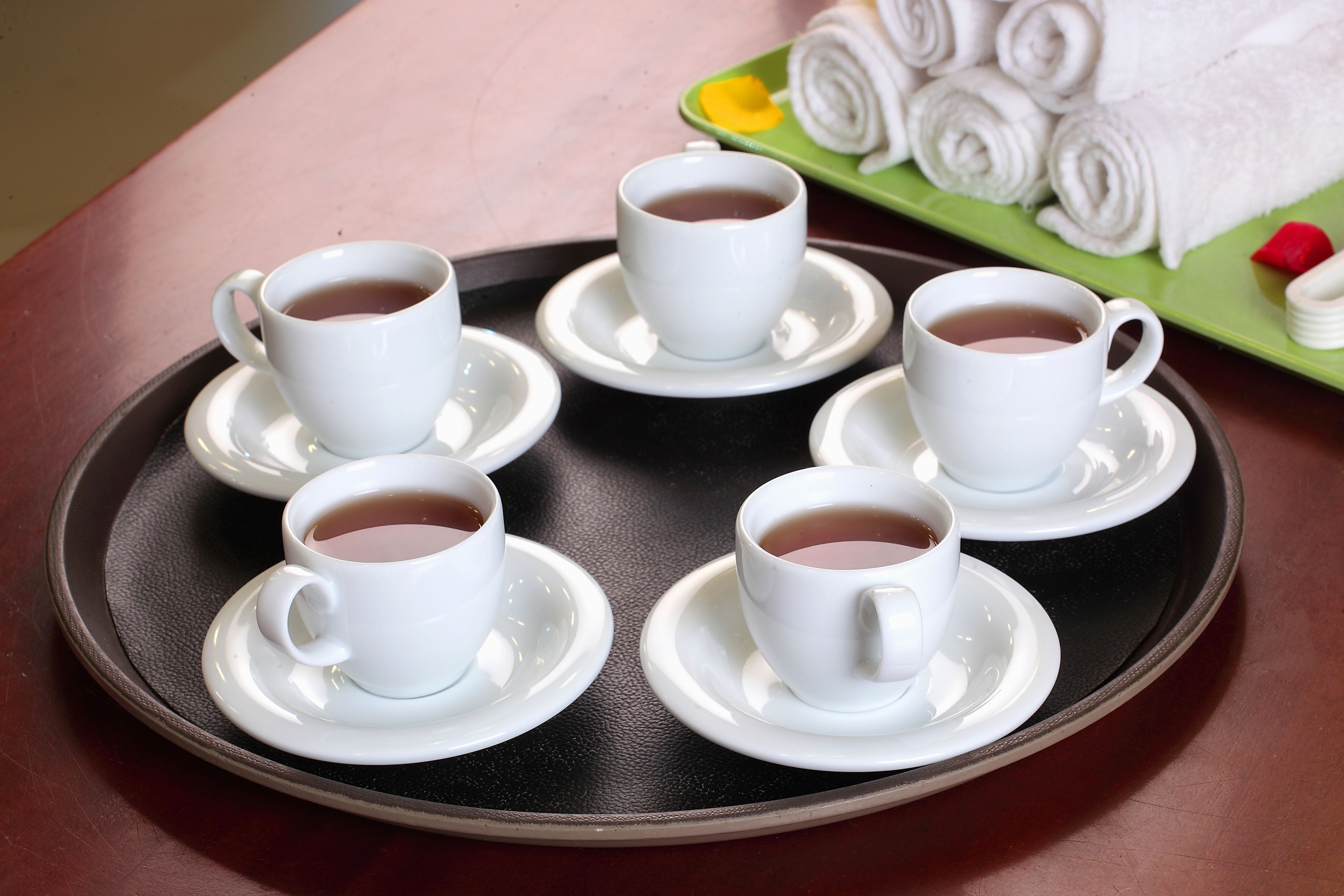 A tray with six white cups of tea on a dark tray, a green tray with rolled white towels, and a red object on the green tray.