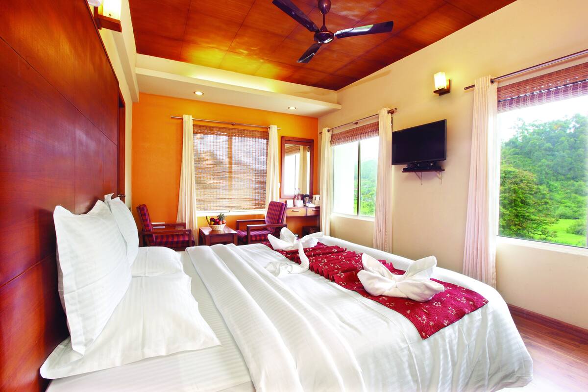 A hotel room with a bed, a desk, a chair, a television, and a window with a view of greenery.