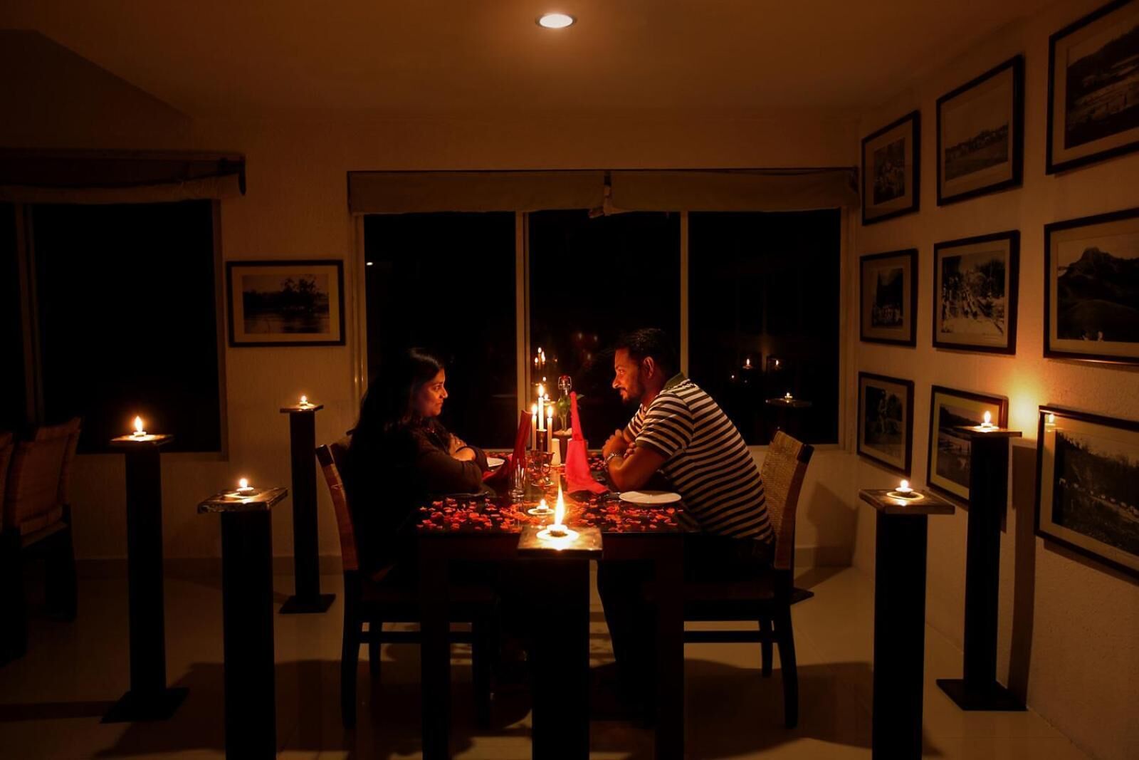 Une salle à manger faiblement éclairée, avec une table dressée pour trois personnes, entourée de bougies et ornée de tableaux encadrés aux murs.