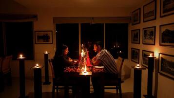 Une salle à manger faiblement éclairée, avec une table dressée pour trois personnes, entourée de bougies et ornée de tableaux encadrés aux murs.