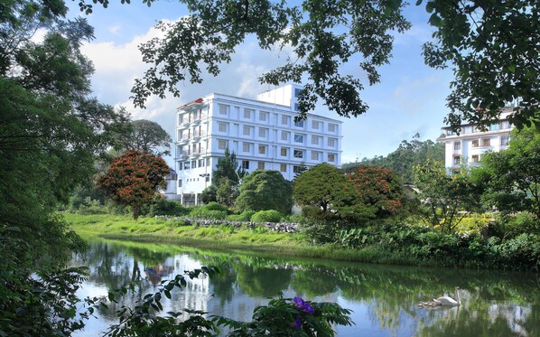 A modern building with multiple floors and balconies, surrounded by lush greenery and a calm water body. - Grand Plaza Munnar (Devikolam)